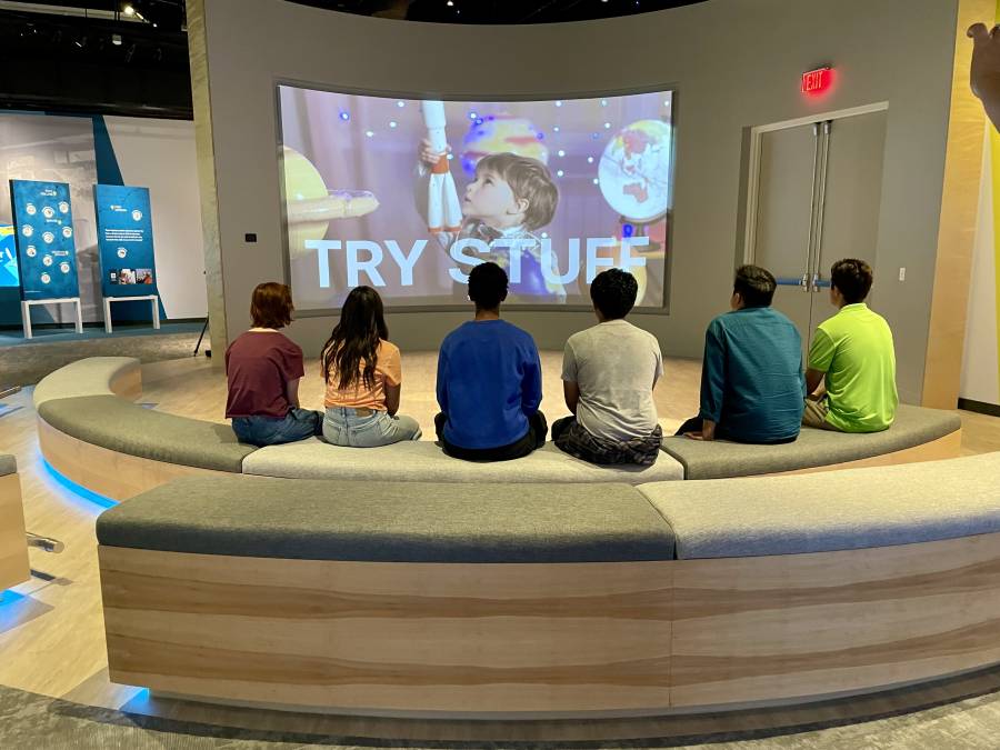 High school students sit in a presentation space where they are shown an introductory video about JA Dream Accelerator